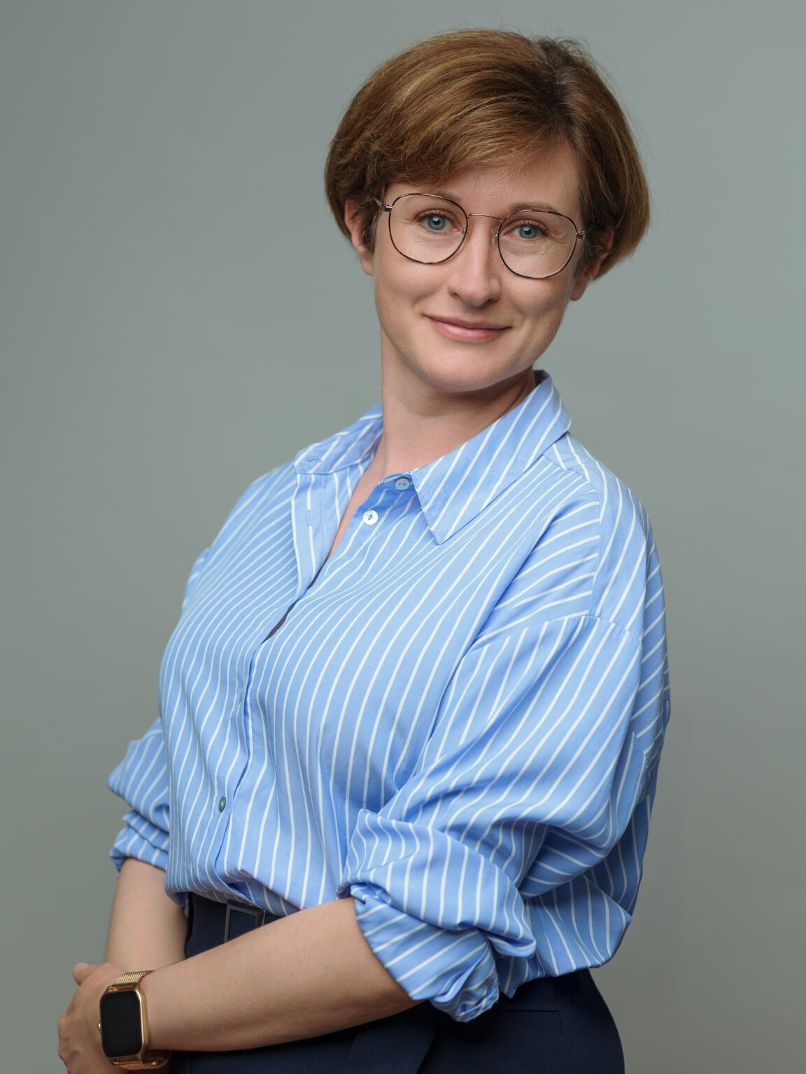 Kobieta o krótkich brązowych włosach, w okularach, niebiesko-białej koszuli w paski i ze smartwatchem, stojąca na jednolitym szarym tle i uśmiechająca się łagodnie do aparatu.