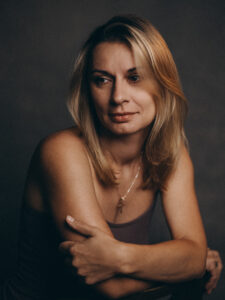 Kobieta o blond włosach, odwracająca wzrok od aparatu, nosząca naszyjnik z małym krzyżykiem.