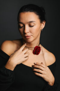 Kobieta ubrana w czarną bluzkę z odkrytymi ramionami i czarny naszyjnik typu choker z zawieszką w kształcie czerwonej róży, dotykająca dłonią swojej szyi.