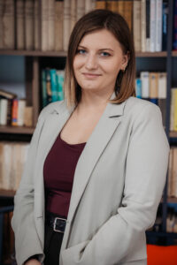 Kobieta w jasnej marynarce i bordowej bluzce, stojąca przed regałem z książkami.