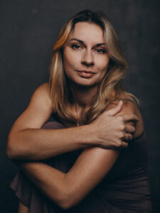 Kobieta o blond włosach sięgających ramion, pozująca ze skrzyżowanymi rękami.